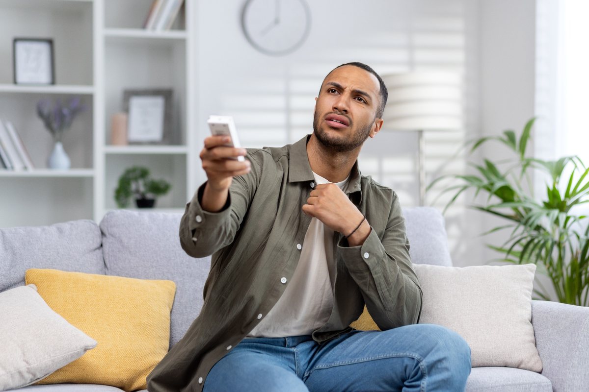 Homem no sofá tentando ligar ar-condicionado com controle remoto sem obter resposta do aparelho
