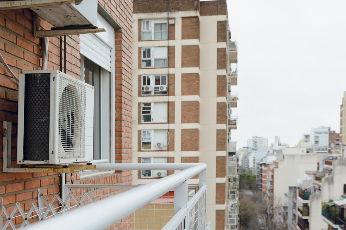 Unidade externa de ar-condicionado fixada à parede de tijolos em varanda de apartamento, com prédios ao fundo.