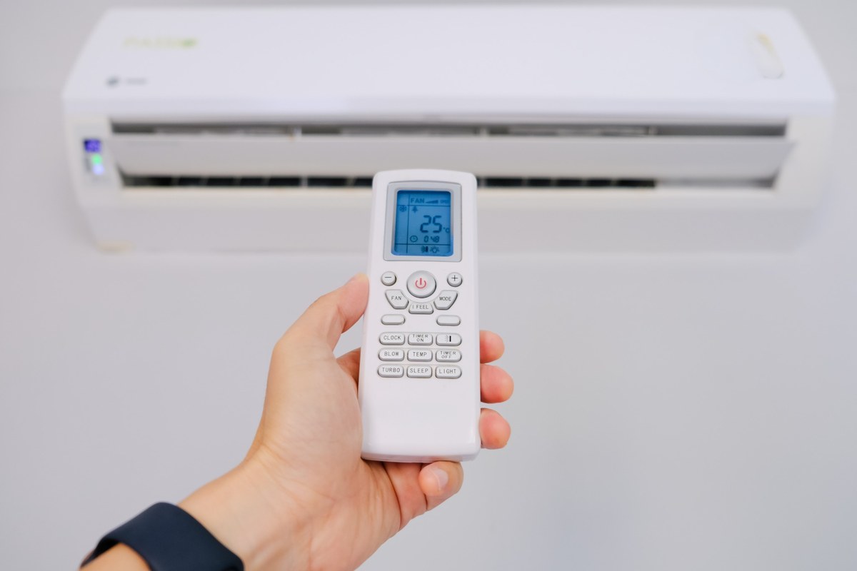 Mão segurando um controle remoto com indicação de 25 °C diante de um ar-condicionado split branco.