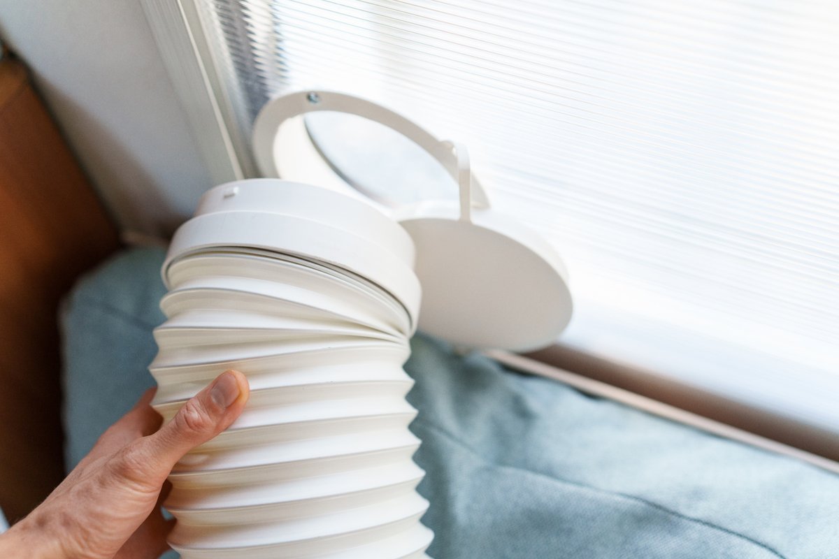 Mão segurando um duto branco de exaustão de ar-condicionado portátil, com adaptador, diante de uma janela com persiana.
